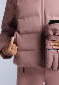 Svjetloružičasta podstavljena jakna s džepom na patentni zatvarač. Crne rukavice od flisa ugurane su u džep jakne radi praktičnosti. Glatka tekstura.