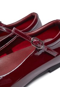 Zapatos de charol rojo con suela plana, punta redondeada y una correa estrecha que presenta una hebilla en tono plateado. Forro interior suave en rojo.