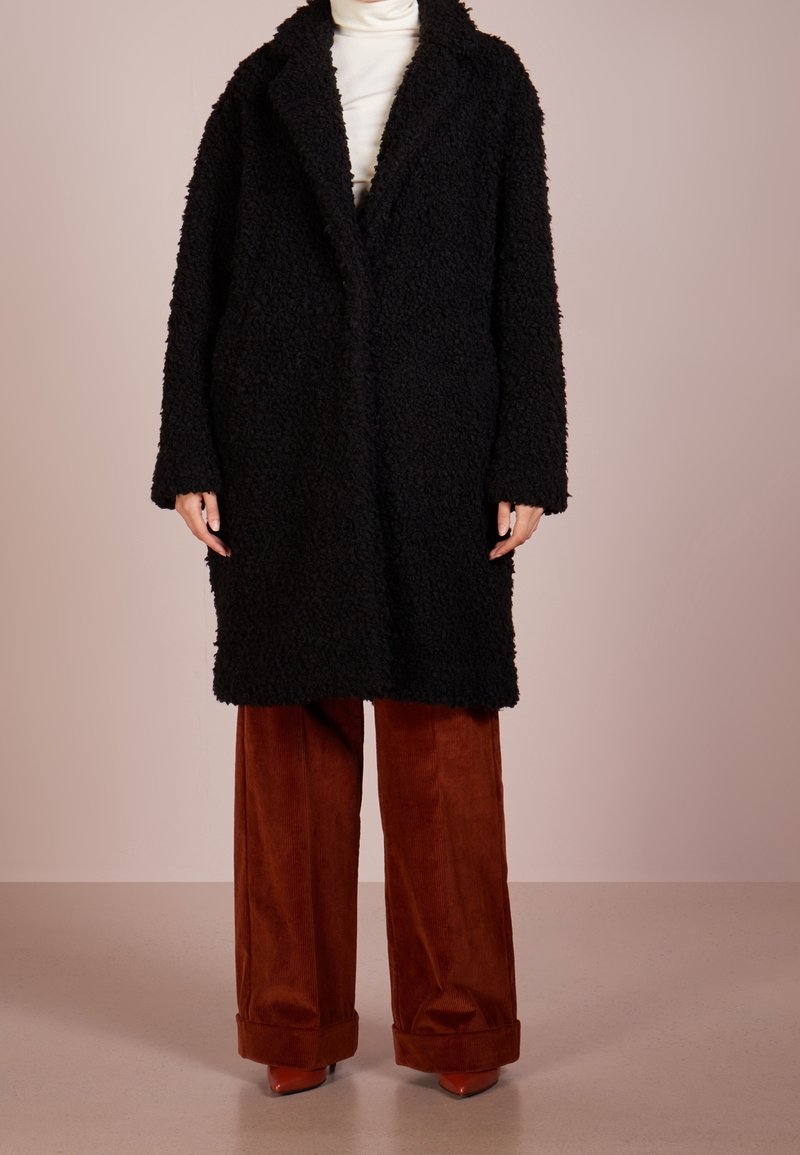 Personne portant un manteau noir texturé par-dessus un col roulé blanc, associé à un pantalon large en velours côtelé couleur rouille et des chaussures marron pointues.