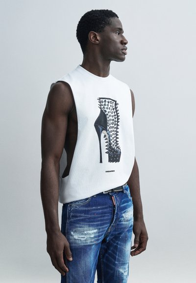 Chemise blanche sans manches avec un graphique de chaussure à talon haut ornée de pointes, associée à un jean bleu déchiré.