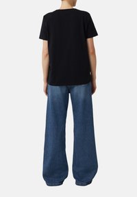 Person wearing a plain black short-sleeve t-shirt and wide-leg blue jeans, standing facing away against a white background.