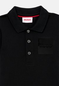 Črna polo majica iz teksturiranega materiala. Ima zapiranje s tremi gumbi ter gumijasti črn logotipni znak na prsih. Rdeča obroba ovratnika.