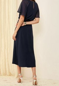 Navy blue dress with short, sheer sleeves and a keyhole back detail. Smooth fabric reaches mid-calf, paired with silver high-heeled sandals.