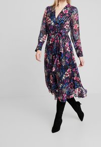 Vestido envolvente floral con mangas largas, que presenta un fondo negro y flores multicolores, acompañado de botas negras hasta la rodilla.
