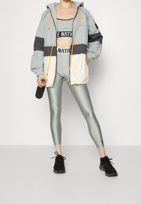 Chaqueta ligera en tonos gris y beige con bloques de color, capucha, puños elásticos y cordones naranjas, combinada con mallas metálicas verdes ajustadas.