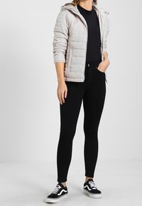 Chaqueta acolchada gris claro con capucha, camiseta negra ajustada, jeans negros skinny y zapatillas negras con suelas y rayas blancas.