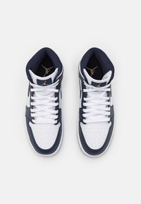 Paire de baskets montantes blanches et bleu marine avec des lacets blancs et des logos Jumpman dorés sur la doublure de la languette bleu marine.