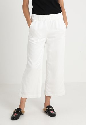 Persoon draagt witte wijde cropped broek met elastische taille en zwarte slip-on schoenen met bont en bloemenborduurwerk. Handen in zakken.