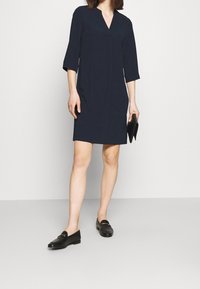 Marinblå V-ringad klänning med trekvartslånga ärmar, i rak skärning och med sidfickor. Bärs med svarta loafers och en kuvertväska.