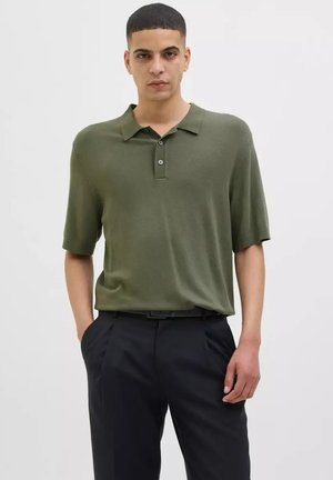 Olivgrünes Poloshirt mit lockerer Passform, kurzen Ärmeln und einer Drei-Knopf-Leiste, kombiniert mit schwarzen Hosen. Glatte Textur.