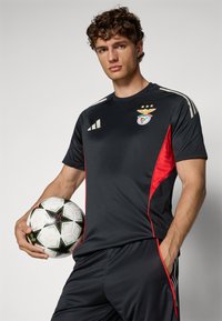 Svart idrottströja med röda accenter och tre vita ränder på ärmarna. Har lagets logotyp och en fotboll med ett stjärnmönster.