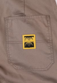 Beige stoffen zak met een gele rechthoekige patch met zwart grafisch ontwerp, tegen een achtergrond van een gladde textuur.
