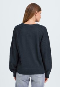 Donker marineblauwe gebreide trui met een gestructureerde achterkant, geribbelde zoom en oversized mouwen. Materiaal lijkt zacht en warm te zijn.