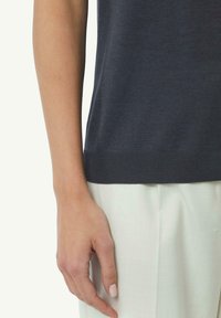 Ciemnoniebieska dzianinowa bluzka z prążkowanym dołem, zestawiona z jasnymi, teksturowanymi spodniami; uwaga skupiona na dłoni opierającej się na tkaninie.