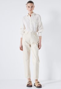 Camisa de botones de color claro y corte holgado con un bolsillo, combinada con pantalones crema de pierna recta y talle alto, y sandalias decoradas.