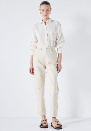 Camisa de botones de color claro y corte holgado con un bolsillo, combinada con pantalones crema de pierna recta y talle alto, y sandalias decoradas.