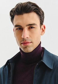 Mężczyzna w ciemnoniebieskiej kurtce na bordowym golfie. Kurtka ma zamek błyskawiczny z przodu i spiczasty kołnierz, z gładką teksturą materiału.