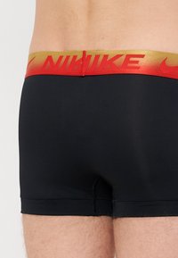 Boxeri atletici negri cu un elastic roșu și auriu, purtând logo-ul "Nike", textură netedă și design ajustat.