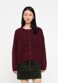 Mulher vestindo um cardigan peludo em bordô com bot�ões e uma saia de veludo cotelê verde escuro com bolsos frontais, em fundo branco liso.