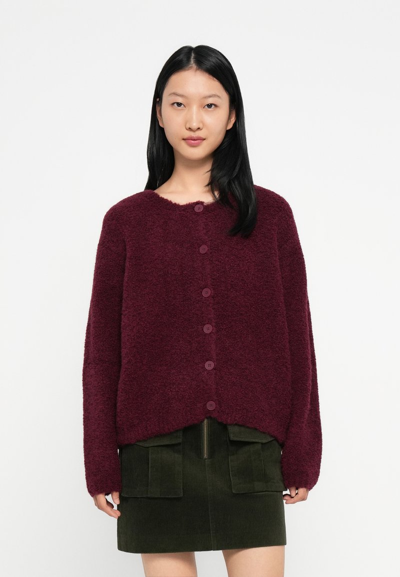 Mulher vestindo um cardigan peludo em bordô com botões e uma saia de veludo cotelê verde escuro com bolsos frontais, em fundo branco liso.