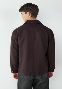 Brązowa, teksturowana kurtka z kołnierzykiem koszulowym, zapięciami na guziki i szwami z tyłu; noszona z ciemnoszarymi spodniami. Pokazuje widok z tyłu.