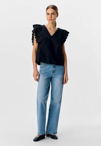 Svart topp med stora volangärmar, V-ringning, i sällskap med ljusblå raka jeans och svarta ballerinas, mot en enfärgad bakgrund.