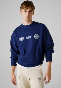 Sweatshirt bleu marine avec col rond, poignets et ourlet côtelés. Présente des logos blancs : "AUTHENTIC RACE GEAR" et "Red Bull RACING."