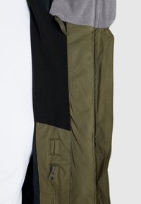 Gros plan sur la manche d'une veste montrant un tissu vert olive, une poche zippée, un panneau noir et une doublure polaire grise au niveau du poignet.