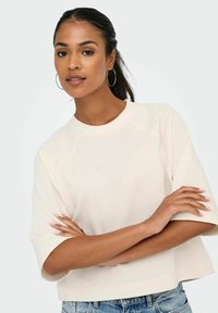 Frau mit dunklem Haar im Pferdeschwanz, die Ohrringe in Form von Ringen trägt, ein cremefarbenes, oversized T-Shirt mit kurzen Ärmeln und blaue Jeans trägt und ihre Arme verschränkt.