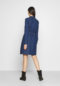 Denim-mekko pitkillä hihoilla, vyötäröltä kiristetty ja liehuva helma. Tumma sininen väri, näkyvät saumat ja shirt collar -yksityiskohta. Yhdistetty nilkkabootsien kanssa.