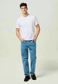 Λευκό βαμβακερό t-shirt και ανοιχτό μπλε τζιν παντελόνι. Το μοντέλο στέκεται χαλαρά με τα χέρια στις τσέπες. Μαύρα παπούτσια ολοκληρώνουν την εμφάνιση.