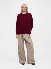 Jeune femme aux longs cheveux blonds portant des lunettes rondes, un pull bordeaux, un pantalon large beige et des chaussures marron, debout devant un fond blanc.