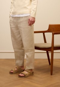 Pantaloni in lino beige con vestibilità ampia, top bianchi a strati e sandali marroni con cinturini regolabili. Sedia di legno sullo sfondo.