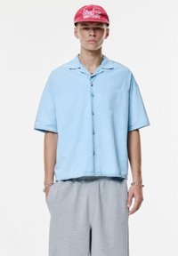 Cămașă cu mâneci scurte, de culoare albastru deschis, cu un croi lejer, combinată cu pantaloni de trening gri. Cămașa are un mic logo brodat pe piept.