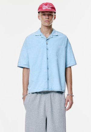Lichtblauw kortemouwen overhemd met een relaxte pasvorm, gecombineerd met grijze sweatpants. Het shirt heeft een klein geborduurd logo op de borst.