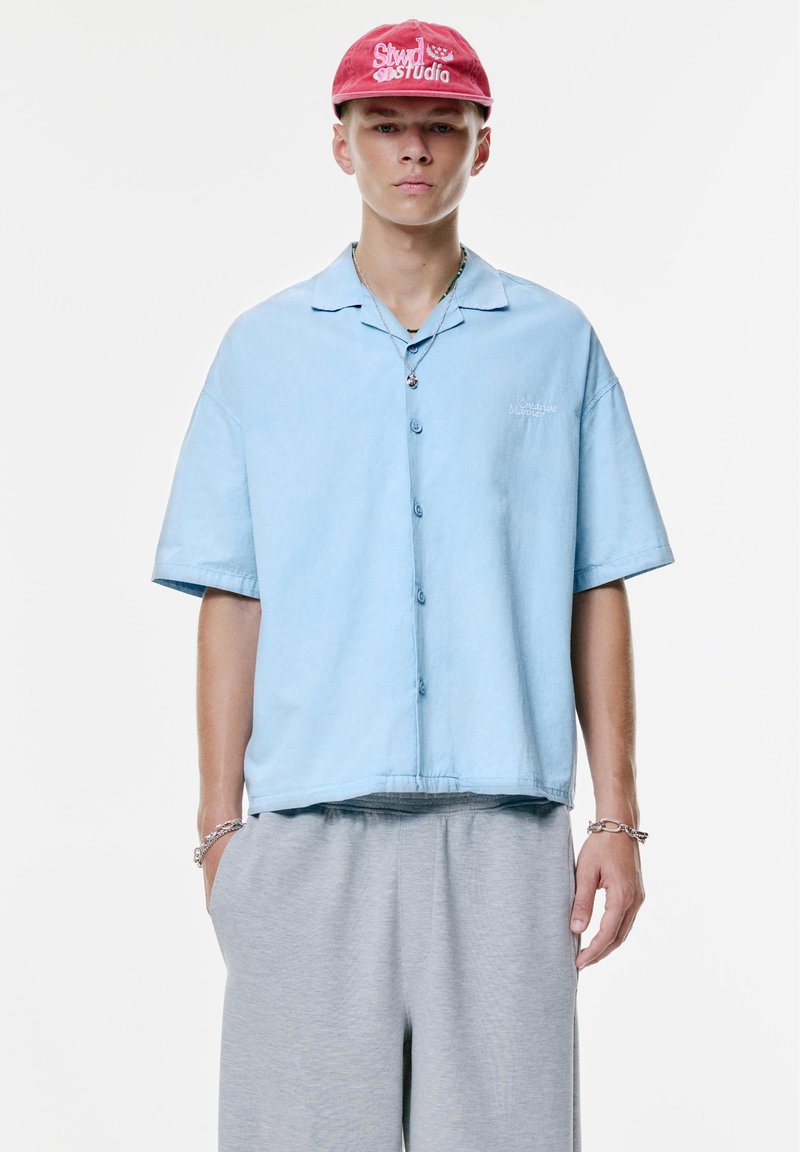 Cămașă cu mâneci scurte, de culoare albastru deschis, cu un croi lejer, combinată cu pantaloni de trening gri. Cămașa are un mic logo brodat pe piept.