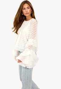 Blouse blanche semi-transparente avec manches à volants, pois texturés et coupe décontractée, associée à un jean droit bleu clair.