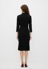 Schwarzes, figurbetontes Kleid mit langen Ärmeln und knielangem Saum. Verfügt über einen hohen Ausschnitt und seitliche Schlitzdetails, kombiniert mit burgunderfarbenen High Heels.