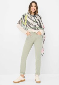 Letvægt, mønstret tørklæde hænger over en beige sweater. Modellen har grønne, tætsiddende bukser og beige sneakers med en lys sål på.