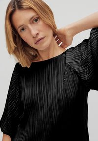 Sort plisseret bluse med en bådformet udskæring og puffede ærmer, der har et tekstureret stof, som skaber lodrette linjer gennem hele blusen.