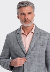Grå strukturerad blazer med ett subtilt rutmönster, med en knappslutning och en bröstficka, bärs över en ljusrosa skjorta.