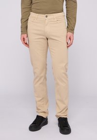 Pantalones de algodón beige con un corte ajustado, que cuentan con bolsillos delanteros y cierre de botón. Llevados con zapatillas negras.