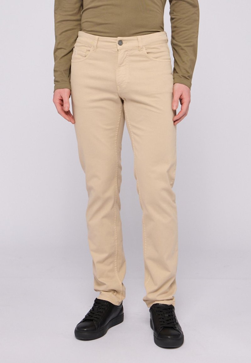 Pantalones de algodón beige con un corte ajustado, que cuentan con bolsillos delanteros y cierre de botón. Llevados con zapatillas negras.