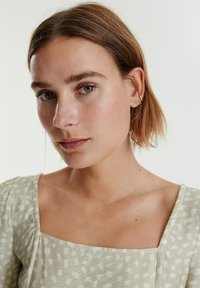 Hellgrünes, gemustertes Oberteil mit quadratischem Ausschnitt und kurzen Ärmeln. Der Stoff hat eine weiche, strukturierte Optik mit weißen Sprenkeln.