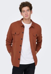 Νέος άνδρας με σγουρά μαλλιά, φοράει ένα καφέ-σκουρό πουκάμισο-ζακέτα με κουμπιά πάνω από ένα λευκό μπλουζάκι και μαύρο παντελόνι, στέκεται μπροστά σε μια απλή φόντο.