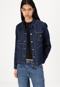 Veste en denim foncé avec des coutures blanches contrastées, dotée d'une poche avant, d'une fermeture à boutons et d'un design court. Associée à un jean bleu clair.