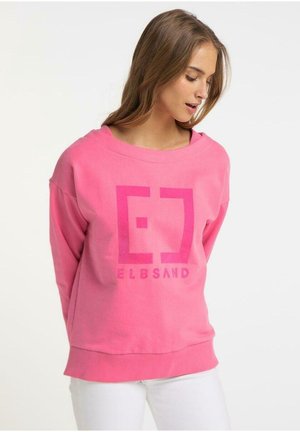 Jeune femme regardant vers le bas, portant un sweat-shirt rose vif avec un logo "ELBSAND" en gros caractères, associé à un pantalon blanc, sur un fond uni.