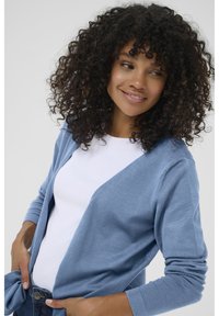 Cardigan azzurro chiaro realizzato in tessuto morbido, con apertura frontale, maniche lunghe e polsini a coste, indossato sopra una maglia bianca.