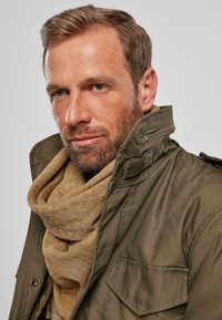 Chaqueta militar de color verde oliva con un cuello alto y cierre de cremallera, superpuesta a una bufanda marrón claro con un patrón sutil.