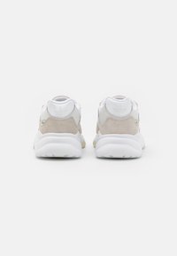 Bakre vy av vita och beige sneakers med en överdel i mocka och mesh, vadderad krage, vit gummisula och präglade logodetaljer.